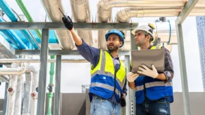 Two engineers discuss plans and use laptop computer while reviewing blueprints on a rooftop.