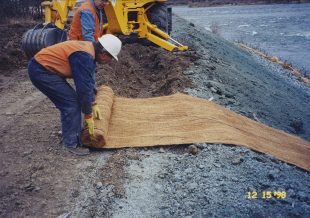 A worker in an orange vest, white hard hat, and blue pants unrolls a large, brown erosion control mat on a sloped, muddy bank