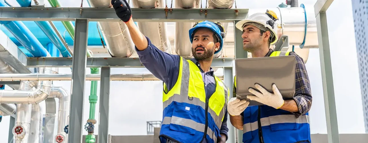 Two engineers discuss plans and use laptop computer while reviewing blueprints on a rooftop.
