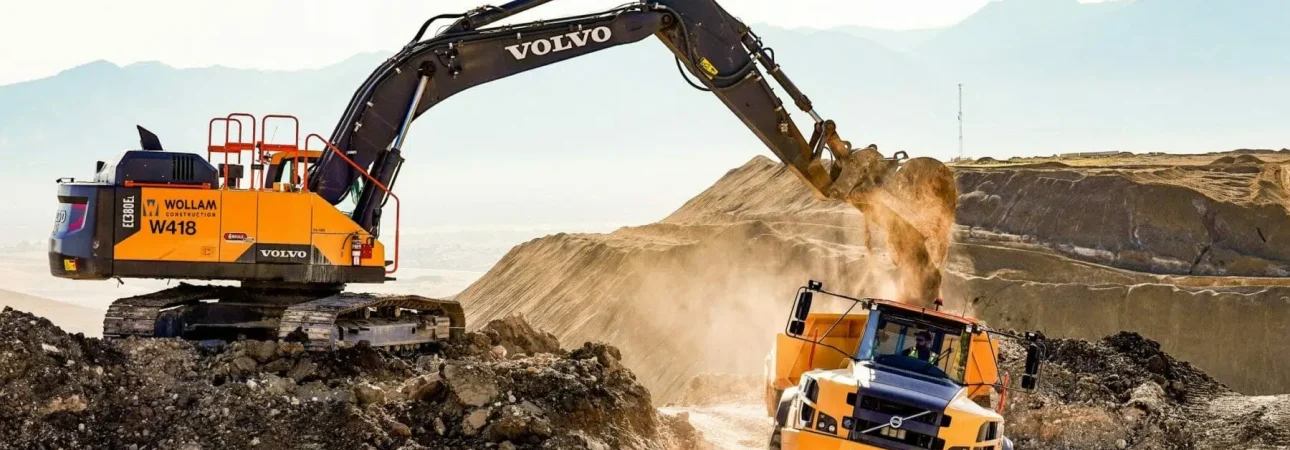 excavator with a large arm digging in the lower left, on a dirt path brown hillside